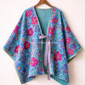 Puede incluir: Un kimono floral turquesa con cuello en V y cierre de lazo. El kimono presenta un vibrante estampado de flores rosas y moradas con detalles en naranja. El texto "Triveni Handicrafts" es visible.