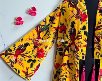 Puede incluir: Un kimono amarillo con estampado floral y de pájaros. La prenda de manga larga presenta detalles rojos, verdes y morados, con borlas rosas. El texto "Triveni handicrafts" es visible en la prenda.