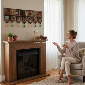 May include: A decorative wall hanging with embroidered panels and tassels hangs above a wooden fireplace. The fireplace has a black insert and is flanked by potted plants and candles. The room is lit by natural light.