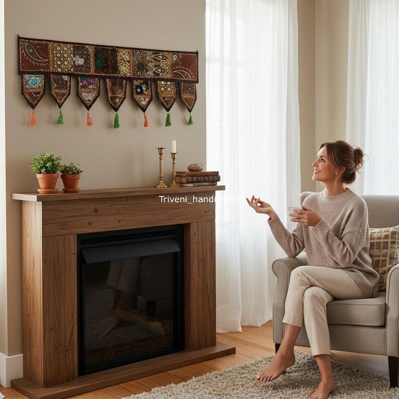 May include: A decorative wall hanging with embroidered panels and tassels hangs above a wooden fireplace. The fireplace has a black insert and is flanked by potted plants and candles. The room is lit by natural light.