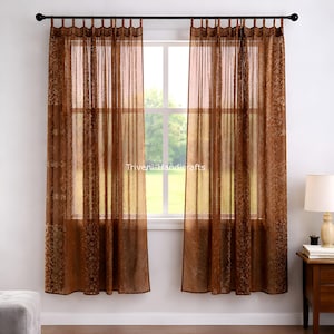 Puede incluir: Dos cortinas marrones transparentes cuelgan de una barra negra frente a una ventana. Las cortinas tienen un estampado floral y son semitransparentes, permitiendo que la luz se filtre. El texto "Triveni Handicrafts" es visible en las cortinas.