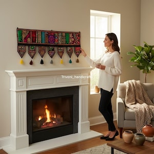 May include: A colorful, embroidered wall hanging with a black border and decorative tassels. The textile features a patchwork design with various patterns and colors, displayed above a white fireplace.