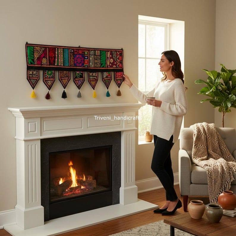 May include: A colorful, embroidered wall hanging with a black border and decorative tassels. The textile features a patchwork design with various patterns and colors, displayed above a white fireplace.