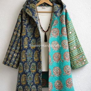 Puede incluir: Una chaqueta larga con capucha y diseño patchwork. La chaqueta presenta un estampado de cachemira azul marino en un lado y un estampado de espirales turquesa en el otro. Las mangas tienen un estampado de rayas con detalles de espejo. Debajo se ven una camiseta blanca y un collar.