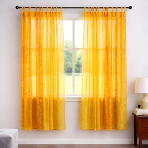 Puede incluir: Un par de cortinas transparentes de color amarillo dorado colgadas frente a una ventana. Las cortinas tienen un patrón texturizado y están suspendidas de una barra negra. El texto "Triveni Handicrafts" es visible en la ventana.
