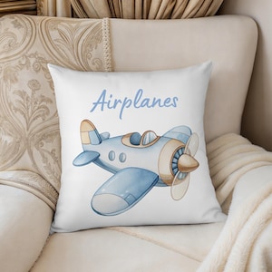 K&ouml;nnte beinhalten: Wei&szlig;es quadratisches Kissen mit einer Aquarellillustration eines blau-braunen Flugzeugs. Das Wort "Airplanes" steht in hellblauer Schrift &uuml;ber dem Flugzeug. Das Kissen liegt auf einem cremefarbenen Stuhl.
