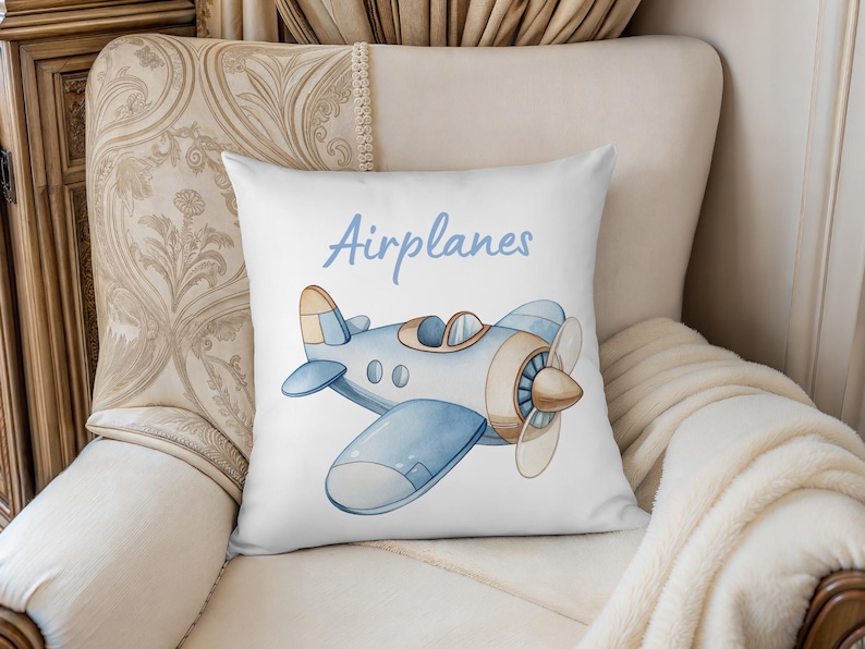 K&ouml;nnte beinhalten: Wei&szlig;es quadratisches Kissen mit einer Aquarellillustration eines blau-braunen Flugzeugs. Das Wort "Airplanes" steht in hellblauer Schrift &uuml;ber dem Flugzeug. Das Kissen liegt auf einem cremefarbenen Stuhl.