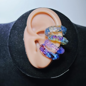 Puede incluir: Cuatro puños de oreja de vidrio de colores se muestran en una oreja de maniquí. Los puños son de varios tonos de azul, morado y amarillo, con una apariencia texturizada y veteada. La oreja es de tono claro y el fondo es negro.