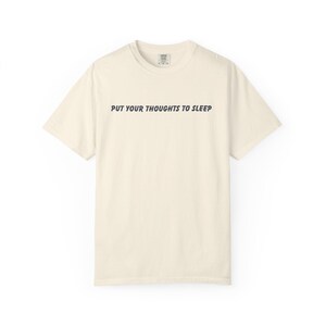 Puede incluir: Camiseta color crema con la frase "PUT YOUR THOUGHTS TO SLEEP" impresa en gris oscuro. La camiseta tiene cuello redondo y mangas cortas. La tela parece suave y cómoda.