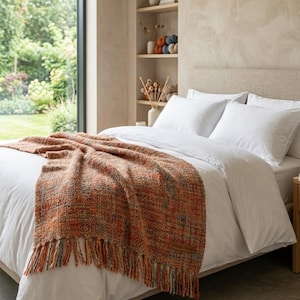 May include: A woven throw blanket in shades of orange, brown, and gray with fringed edges, draped over a bed with white linens. The blanket has a textured appearance and a rectangular shape.