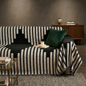 May include: A black and white throw blanket with a repeating vertical leaf-like pattern draped over a sofa. A solid dark green velvet square throw pillow rests on the blanket. A small glass candle sits on a book atop the sofa cushion.