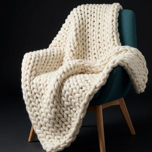 Puede incluir: Una manta de punto grueso color crema sobre un sillón verde azulado con patas de madera. El hilo grueso de la manta crea una apariencia texturizada y acogedora, ideal para la decoración del hogar.