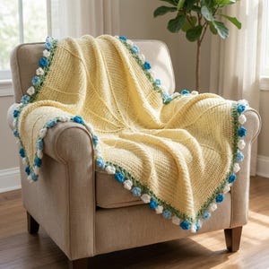 May include: A yellow crocheted blanket with a textured pattern is draped over a beige armchair. The blanket is trimmed with a border of blue, white, and green pom-poms, adding a decorative touch to the cozy home decor item.