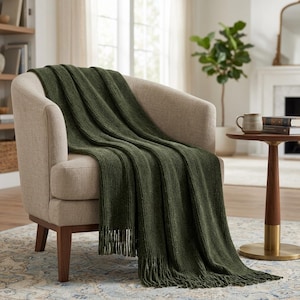 May include: An olive green knitted throw blanket with fringe detail draped over a beige armchair. The blanket has a ribbed texture and is made of a soft material. The chair has wooden legs and is placed on a patterned rug.