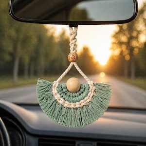Puede incluir: Un adorno de macramé para coche colgado de un espejo retrovisor. El adorno presenta un diseño en forma de abanico con flecos verde salvia, una cuenta de madera natural y cuerda de color crema. El fondo muestra una carretera y árboles.