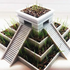 Puede incluir: Una maceta gris impresa en 3D con forma de pirámide con múltiples niveles y escaleras. Cada nivel contiene tierra y hierba verde brotando. La maceta está diseñada para parecerse a un templo o estructura antigua.