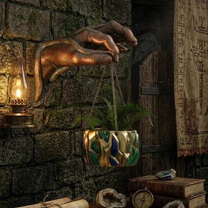 May include: A bronze hand sculpture holds a hanging planter with a geometric design of gold, green, and blue. The planter contains green ferns. An oil lamp and a stone wall are in the background. The scene evokes a fantasy or historical theme.