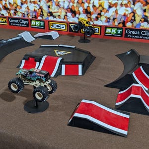 May include: A miniature monster truck with a colorful design sits on a stand, surrounded by black, red, and white ramps and obstacles. The scene is set against a backdrop of a crowd and advertising banners, creating a miniature monster truck rally.