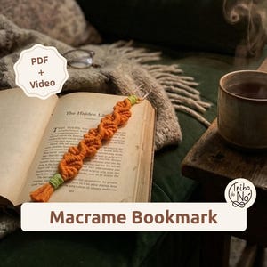 Puede incluir: Un marcapáginas de macramé naranja con un diseño de cordón trenzado, detalles en verde claro en ambos extremos y una pequeña borla en la parte inferior. El marcapáginas está sobre un libro abierto. La imagen incluye el texto PDF + Video y Macrame Bookmark.