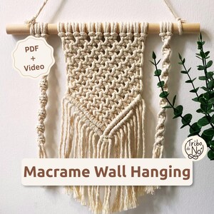 Pode incluir: Uma tapeçaria de macramé cor de creme suspensa num bastão de madeira com um cordão. A peça apresenta um padrão central de nós com franjas e dois fios laterais. O texto na imagem diz PDF + Video e Macrame Wall Hanging.