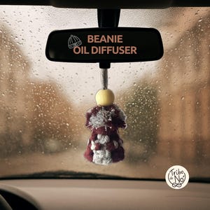Puede incluir: Un espejo retrovisor de coche con el texto "Beanie Oil Diffuser" en rosa. Debajo, cuelga un pequeño difusor de aceite hecho a mano con una cuenta de madera amarilla y un diseño de ganchillo en rojo, blanco y gris. El fondo muestra un día lluvioso.