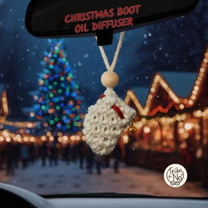 Può includere: Un diffusore di olio a forma di stivale natalizio all'uncinetto color crema, appeso allo specchietto retrovisore di un'auto. Lo stivale ha un accento rosso e una piccola campana dorata. Il testo "CHRISTMAS BOOT OIL DIFFUSER" è sopra lo specchietto. Un albero di Natale e luci festive sullo sfondo.