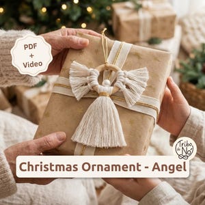 May include: A macrame angel Christmas ornament featuring a light wood ring, off-white cotton cord wings, a central white bead, and a long tassel skirt with gold thread accents. The ornament is displayed on a gift box with the text PDF + Video and Christmas Ornament - Angel.