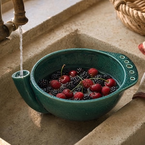 May include: A deep teal ceramic berry bowl with a side spout and a handle featuring five small circular indentations. The bowl is filled with water, fresh red cherries, and dark blackberries, positioned under a running kitchen faucet stream.