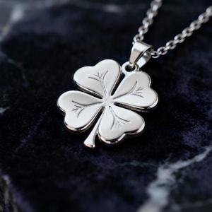 Puede incluir: Un collar con colgante plateado con un diseño de trébol de cuatro hojas. El trébol tiene vetas detalladas en las hojas y cuelga de una cadena fina. El collar se muestra sobre un fondo oscuro y texturizado.