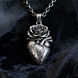 Puede incluir: Collar con colgante de plata que presenta un diseño detallado de corazón anatómico con una rosa en la parte superior. El colgante está suspendido de una cadena de plata. El collar se muestra sobre un fondo oscuro y texturizado.