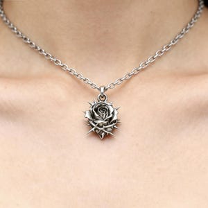 Puede incluir: Un collar plateado con un colgante en forma de rosa rodeada de espinas. El colgante cuelga de una cadena. La rosa tiene p&eacute;talos detallados y est&aacute; rodeada de afiladas espinas.