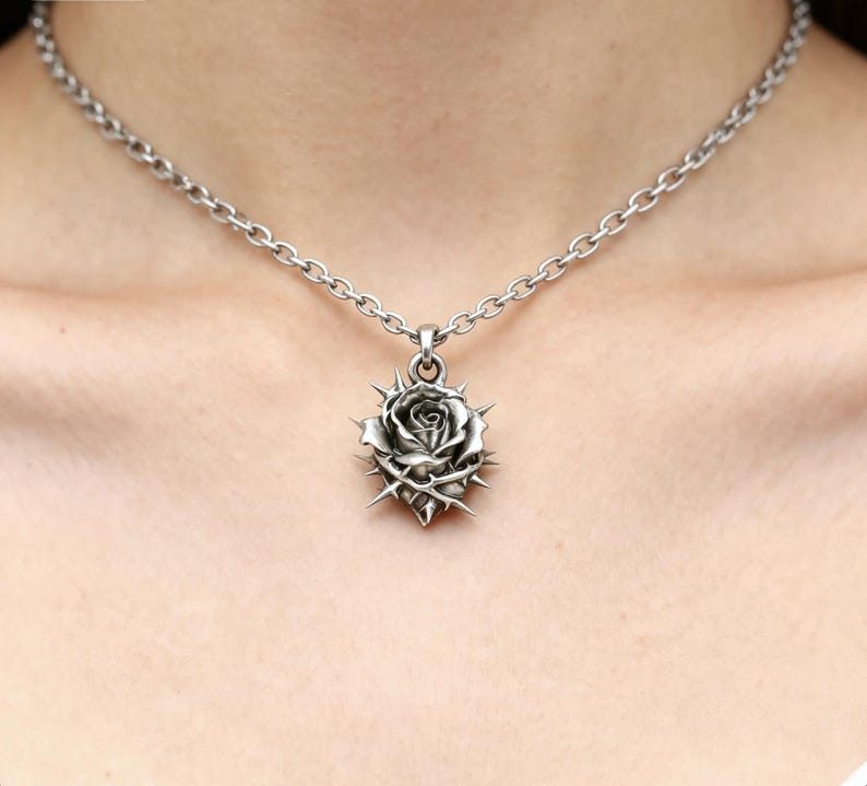 Puede incluir: Un collar plateado con un colgante en forma de rosa rodeada de espinas. El colgante cuelga de una cadena. La rosa tiene p&eacute;talos detallados y est&aacute; rodeada de afiladas espinas.