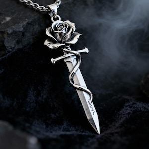 Può includere: Una collana con pendente in argento con un design a rosa e pugnale. La rosa è posizionata sopra il pugnale, con una vite che si avvolge attorno alla lama. Il pendente è appeso a una catena d'argento, creando un'estetica gotica o romantica.