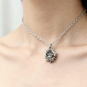 Puede incluir: Collar plateado con un colgante de rosa con espinas. El colgante es detallado, mostrando los p&eacute;talos de la rosa y las espinas. La cadena es de dise&ntilde;o simple, creando una est&eacute;tica g&oacute;tica. El collar se lleva alrededor del cuello.