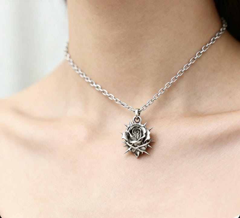 Puede incluir: Collar plateado con un colgante de rosa con espinas. El colgante es detallado, mostrando los p&eacute;talos de la rosa y las espinas. La cadena es de dise&ntilde;o simple, creando una est&eacute;tica g&oacute;tica. El collar se lleva alrededor del cuello.