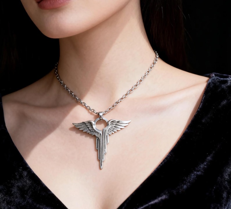 K&ouml;nnte beinhalten: Silberfarbene Anh&auml;ngerkette mit einem stilisierten Fl&uuml;geldesign. Der Anh&auml;nger hat einen zentralen Ring mit detaillierten Fl&uuml;geln und vertikalen Linien. Die Halskette hat eine passende silberne Farbe.