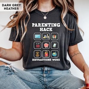 Puede incluir: Camiseta gris oscuro jaspeado con el texto "PARENTING HACK" y "DISTRACTIONS WORK". La camiseta presenta una cuadrícula de iconos ilustrados, incluyendo un televisor, juguetes, lápices de colores, una radio, un libro, un teléfono, un osito de peluche, snacks y un cuenco.