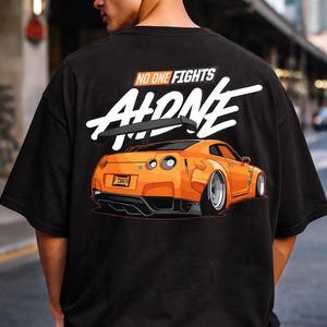 May include: Black t-shirt featuring an orange sports car graphic with the text "NO ONE FIGHTS ALONE" in white. The car design includes details like a spoiler and custom wheels. The shirt is a casual wear item.