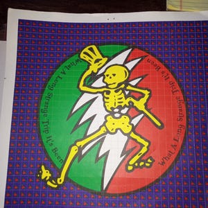 May include: A colorful graphic print featuring a yellow skeleton figure with a top hat and cane, set against a red, green, and white background. The text "What A Long Strange Trip It's Been" encircles the central image. The background is a grid of small red shapes on a purple field.