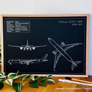 Pode incluir: Impressão emoldurada com um desenho de linha branca de um Airbus A350 XWB A350-900 em um fundo preto. A impressão inclui especificações técnicas e três vistas diferentes da aeronave. A moldura é feita de madeira clara.
