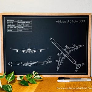 May include: A framed black and white blueprint of an Airbus A340-600 airplane. The print includes technical specifications and multiple views of the aircraft. The frame is made of light-colored wood. The text "Airbus A340-600" is visible.