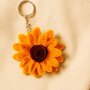May include: A yellow and brown sunflower keychain with a silver keyring and chain. The sunflower has a textured, plush appearance and is set against a beige background, suggesting a tote bag.