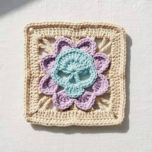 May include: A square, crocheted piece featuring a skull design. The skull is light blue, surrounded by a lavender flower, and set against a beige square background. The piece is handmade, showcasing a unique, handcrafted aesthetic.