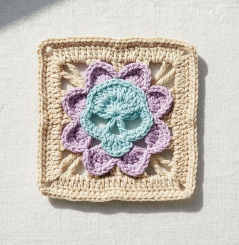 May include: A square, crocheted piece featuring a skull design. The skull is light blue, surrounded by a lavender flower, and set against a beige square background. The piece is handmade, showcasing a unique, handcrafted aesthetic.