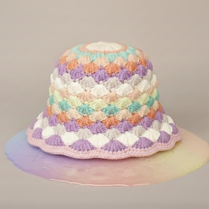 Könnte beinhalten: Ein mehrfarbiger gehäkelter Fischerhut. Der Hut hat ein Muschelmuster in Pastelltönen von Lila, Weiß, Pfirsich, Grün und Blau. Der Rand ist mit einem lavendelfarbenen Häkelstich versehen. Ein modisches Accessoire.