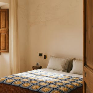 May include: A bed with a handmade crochet blanket. The blanket features a repeating pattern of yellow suns within blue squares. The bed has white pillows and a brown bedspread. The room has a rustic aesthetic with a wooden shutter and door.