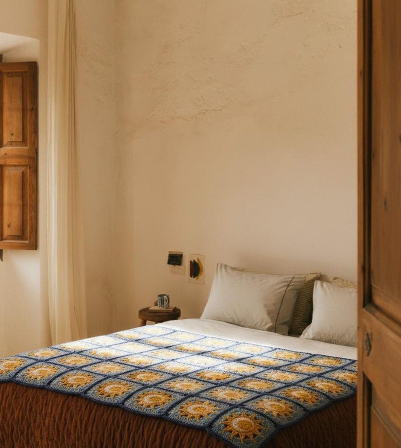 May include: A bed with a handmade crochet blanket. The blanket features a repeating pattern of yellow suns within blue squares. The bed has white pillows and a brown bedspread. The room has a rustic aesthetic with a wooden shutter and door.