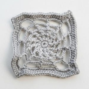 May include: A square, crocheted doily in shades of gray. The center features a spiral design, surrounded by an open, web-like pattern. The edges are finished with a solid, textured border. The handmade piece is displayed on a white surface.