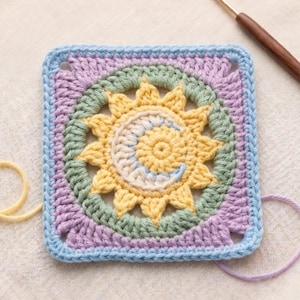 May include: A square, handmade crochet piece featuring a sun and moon design. The sun is yellow with a white and blue crescent moon in the center. The piece is bordered by light blue, lavender, and green yarn.