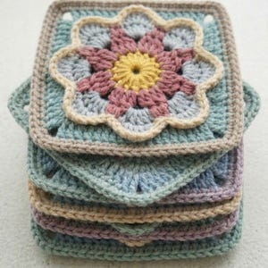 Könnte beinhalten: Ein Stapel handgefertigter Häkelquadrate in Pastellfarben. Jedes Quadrat hat ein zentrales Blumendesign mit gelben, rosa und grauen Blütenblättern, umgeben von einem beigen Rand. Die Quadrate sind in Blau-, Grün- und Lilatönen gehalten.